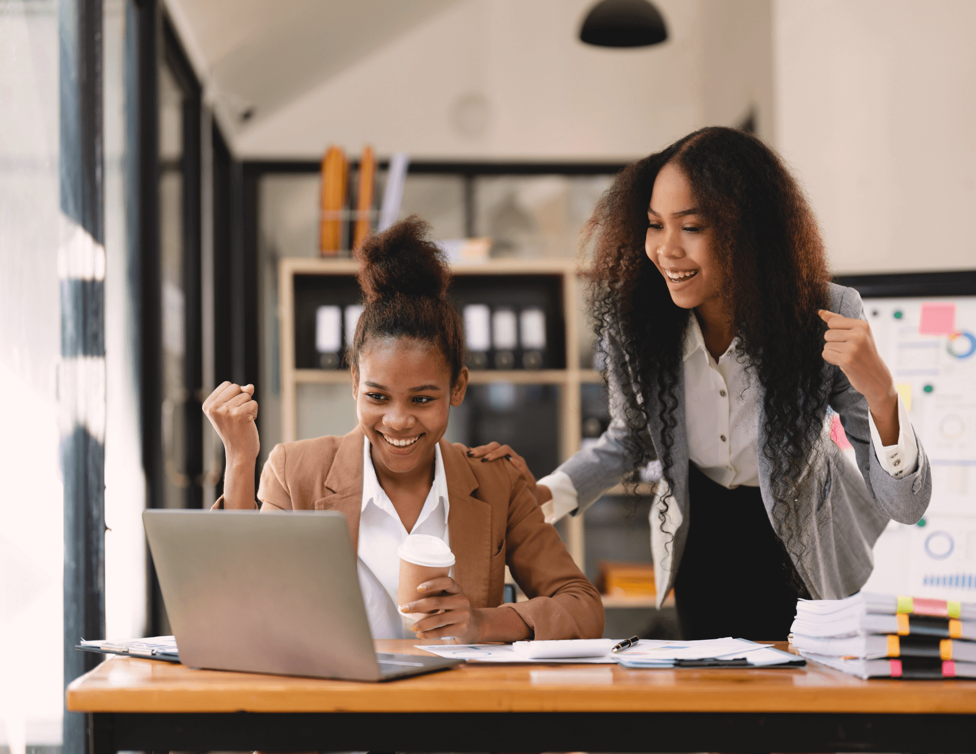 Two professional women in an office celebrating a career win, with one sitting at a desk holding a coffee cup and raising her fist in excitement, and the other smiling with encouragement.